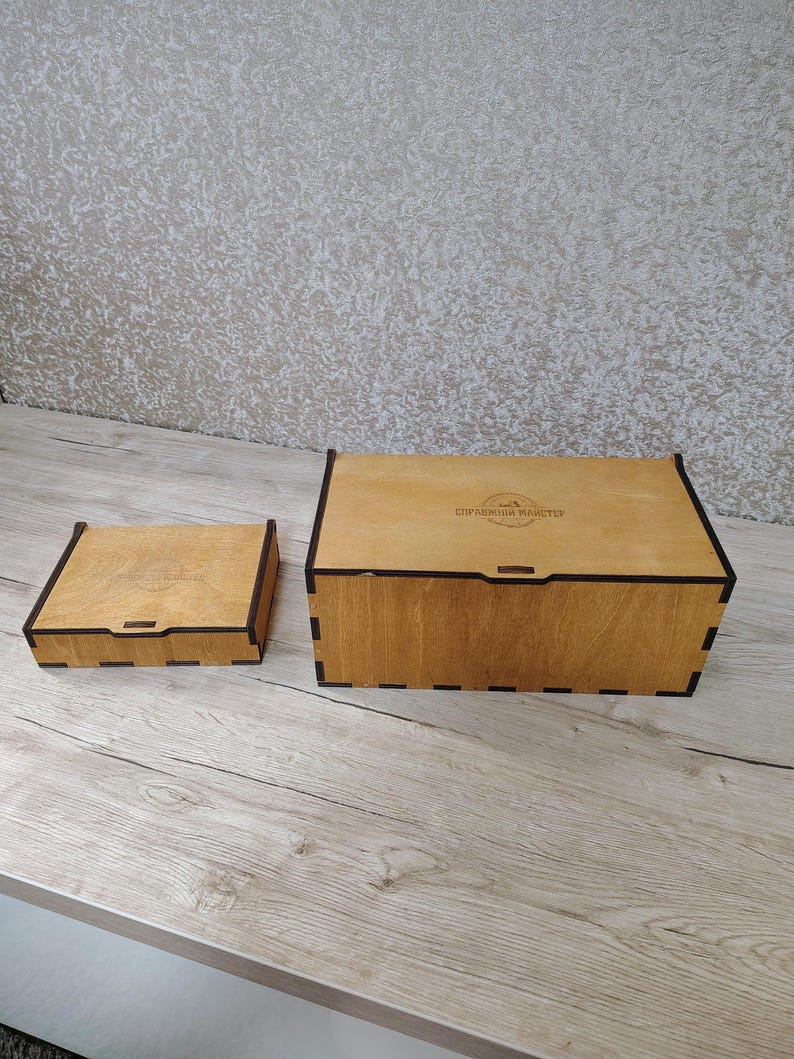 May include: Two wooden boxes with black trim. The larger box is rectangular, and the smaller box is square. Both boxes are made of light-colored wood and have a hinged lid. The boxes are sitting on a light-colored wooden surface.