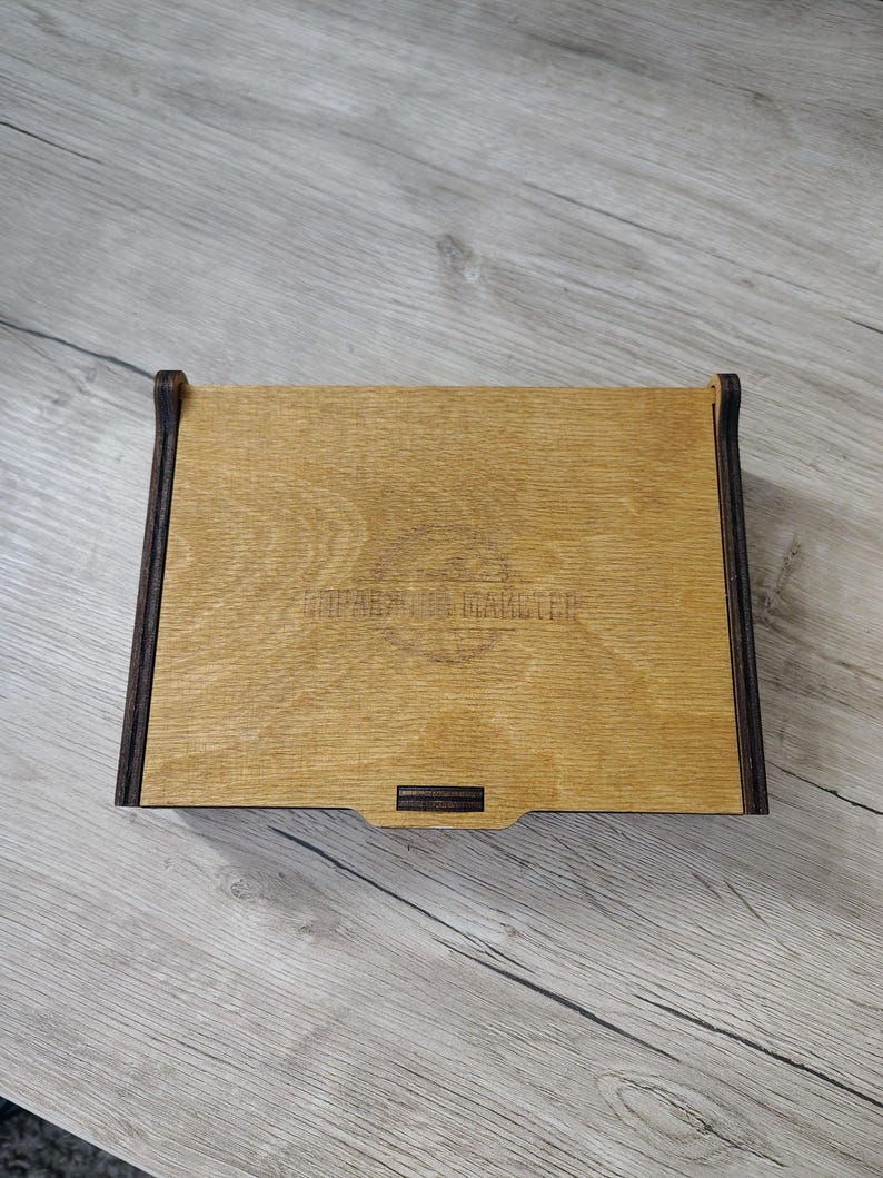 May include: A rectangular wooden box with a light brown top and dark brown edges. The box has a small rectangular indentation on the front. The text "ПРАВИЛА МАСТЕРА" is visible on the top.