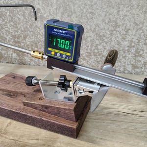 May include: A knife sharpening system with a digital inclinometer displaying 17.00 degrees. The system includes a wooden base, metal components, and a sharpening guide. The digital display is blue and yellow.