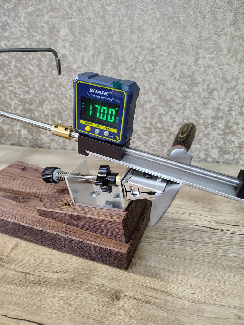 May include: A knife sharpening system with a digital inclinometer displaying 17.00 degrees. The system includes a wooden base, metal components, and a sharpening guide. The digital display is blue and yellow.