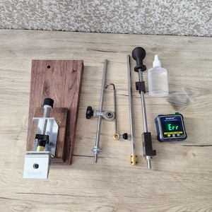 May include: A collection of metal and wood tools, including a wooden base with the text "Sytools", various metal rods, a small bottle, and a digital level displaying "Err". The tools are arranged on a light wood surface.