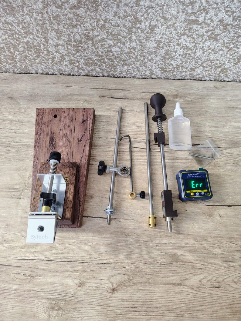 May include: A collection of metal and wood tools, including a wooden base with the text "Sytools", various metal rods, a small bottle, and a digital level displaying "Err". The tools are arranged on a light wood surface.