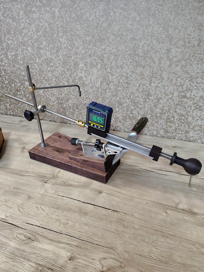 May include: A knife sharpening system with a wooden base, metal components, and a digital angle gauge displaying "16.95". The system includes a black handle and a black knob for adjustments. The background is a textured, neutral color.