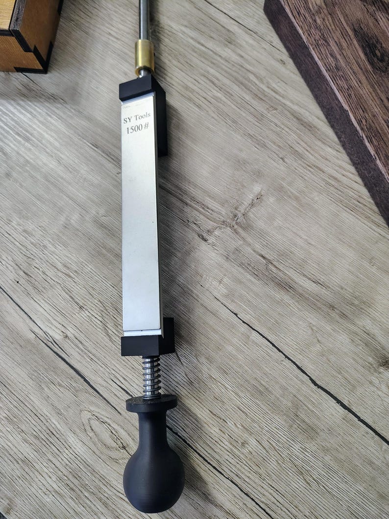 May include: A silver and black knife sharpener with the text "SY Tools 1500 #" printed on the blade. The sharpener has a black handle and a silver blade. The tool is designed for sharpening knives.