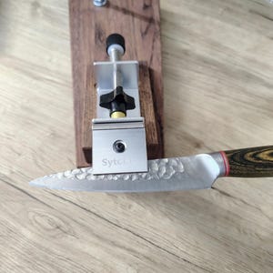 May include: A knife sharpening system with a stainless steel blade and a wooden handle. The blade has a textured surface and the handle has a wood grain pattern. The system includes a metal clamp and a wooden base.