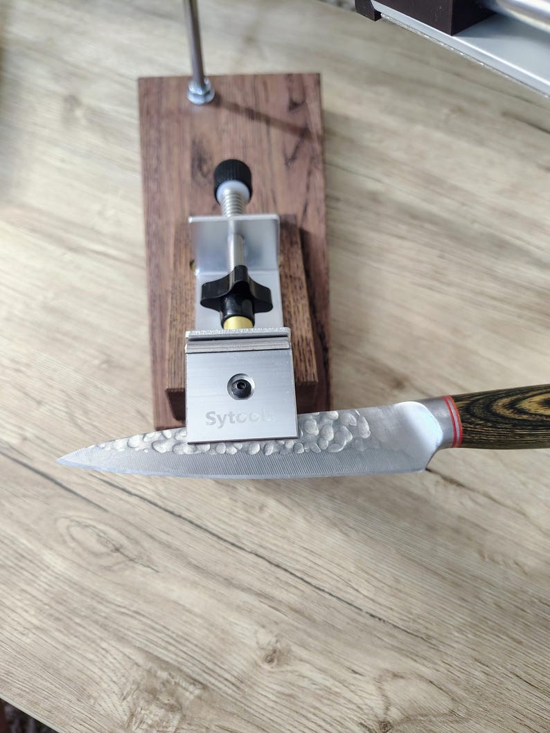 May include: A knife sharpening system with a stainless steel blade and a wooden handle. The blade has a textured surface and the handle has a wood grain pattern. The system includes a metal clamp and a wooden base.