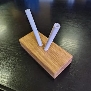 May include: A wooden desk organizer with two white cylindrical pen holders. The organizer is made of light brown wood and sits on a dark surface. The pen holders are angled and appear to be made of a smooth, matte material.