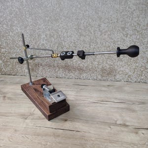 May include: A knife sharpening system with a wooden base and metal components. The system includes a clamp, adjustable arm, and a handle. The base is made of dark brown wood, and the metal parts are silver and black.