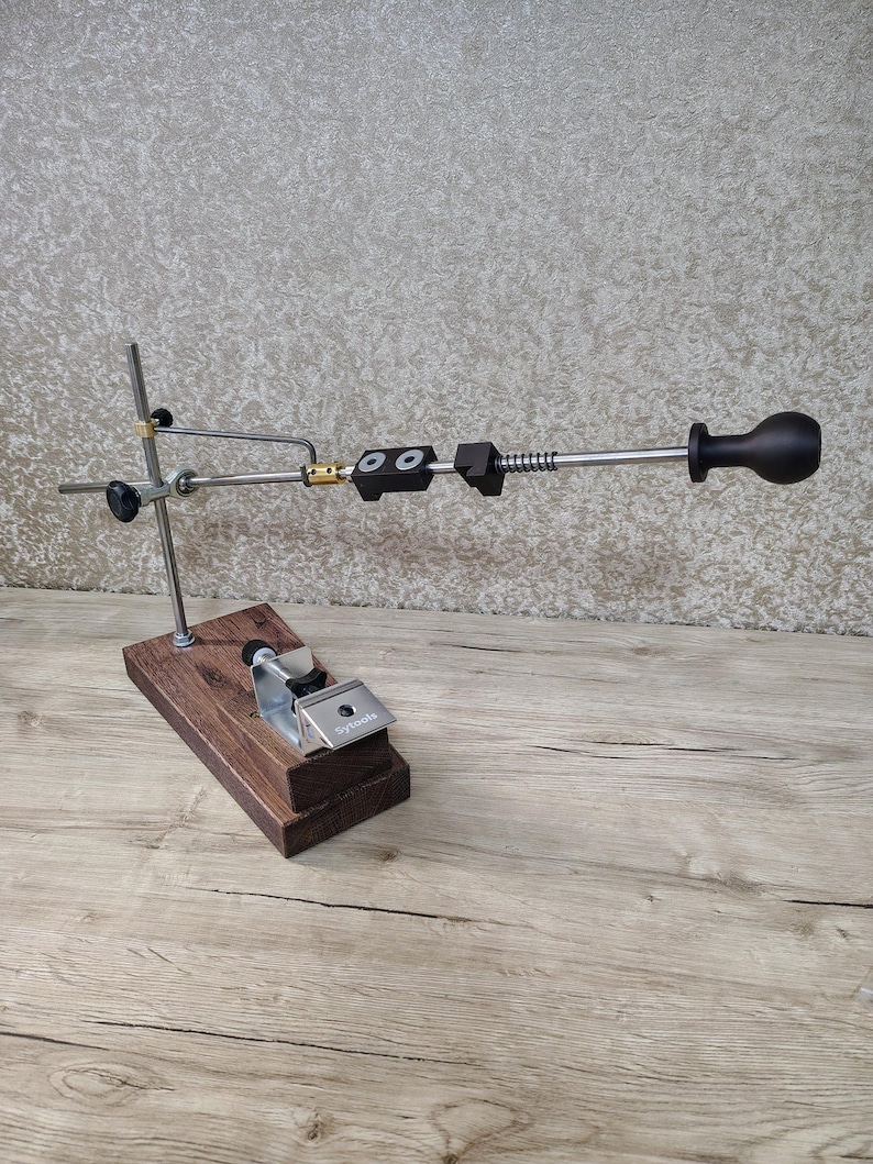 May include: A knife sharpening system with a wooden base and metal components. The system includes a clamp, adjustable arm, and a handle. The base is made of dark brown wood, and the metal parts are silver and black.