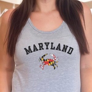 May include: Gray tank top with a black and white Maryland crab graphic. The crab is holding the Maryland state flag. The text "MARYLAND" is printed in black above the crab.