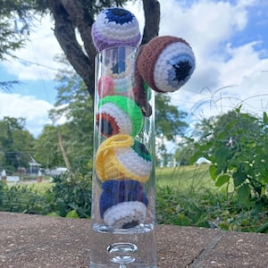 Könnte beinhalten: Eine klare Glasvase gefüllt mit mehreren gehäkelten Augäpfeln in verschiedenen Farben, darunter Lila, Braun, Grün, Gelb und Blau. Die Augäpfel haben weiße und schwarze Details, und einige haben rote Akzente. Die Vase steht auf einer Steinoberfläche mit einem verschwommenen Hintergrund aus Bäumen und Himmel.