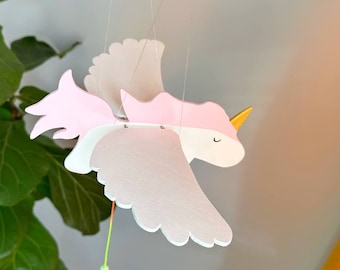 May include: A pink and white wooden unicorn mobile with gold accents. The unicorn has wings and a green bead hanging from its tail.