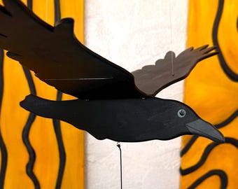 May include: A black wooden crow mobile with outstretched wings, suspended in front of two yellow and black abstract art panels. The crow has a detailed beak and eye, creating a striking contrast against the background.