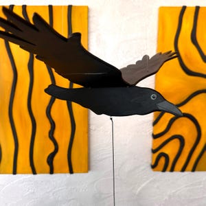 Könnte beinhalten: Ein schwarzes Holz-Krähen-Mobile mit ausgebreiteten Flügeln, aufgehängt vor zwei gelb-schwarzen abstrakten Kunsttafeln. Die Krähe hat einen detaillierten Schnabel und ein Auge, und die Flügel sind leicht angewinkelt.