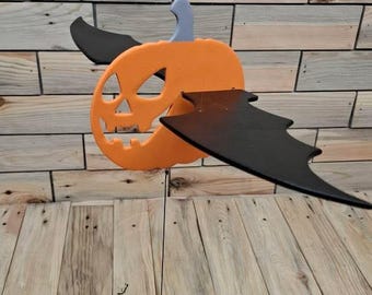 May include: Orange pumpkin decoration with a carved face and black bat wings. The pumpkin has a gray stem and is suspended by a string with a wooden bead. The background is a wooden plank wall.