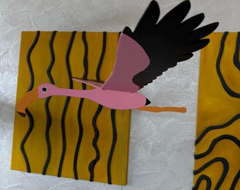May include: A pink flamingo mobile with black and pink wings, an orange beak, and a green string. The flamingo is in flight against a background of yellow squares with black wavy lines.