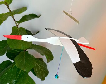 May include: A white and black wooden stork mobile with a red beak and a blue and green hanging bead. The mobile is suspended from a wooden dowel.