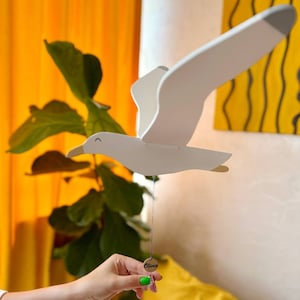 May include: White wooden seagull mobile with a silver tag that says "Olivia". The mobile is hanging from a string and is being held by a hand with green nail polish.
