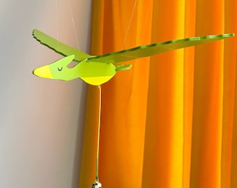May include: A wooden, lime green pterodactyl mobile with a yellow beak and a small, patterned ball hanging below. The mobile is suspended by thin strings against a backdrop of an orange curtain and a white wall.