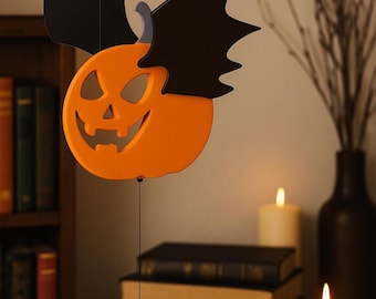 May include: Orange pumpkin decoration with black bat wings, hanging from a thin string. The pumpkin has a carved face with a smiling expression. The background includes a bookshelf, candles, and a vase.