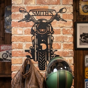 Può includere: Decorazione da parete a forma di moto in metallo nero con il nome "SMITH'S" sopra il manubrio. Una giacca di pelle marrone è appesa sotto e un casco da moto verde è montato sul muro di mattoni. Sono visibili anche insegne di moto vintage.