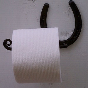 May include: A black metal horseshoe toilet paper holder with a white roll of toilet paper. The holder is mounted to a white wall.