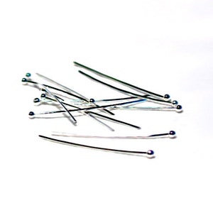 May include: A collection of silver-colored head pins with small, round, silver-colored heads. The pins are scattered on a white surface, with some overlapping. The pins are straight and have a smooth, reflective surface. These pins are used for jewelry making and crafting.