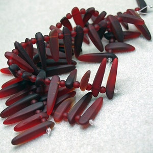 May include: A strand of dark red and amber glass beads in a teardrop shape. The beads are strung on a clear thread and are arranged in a loose, flowing pattern.