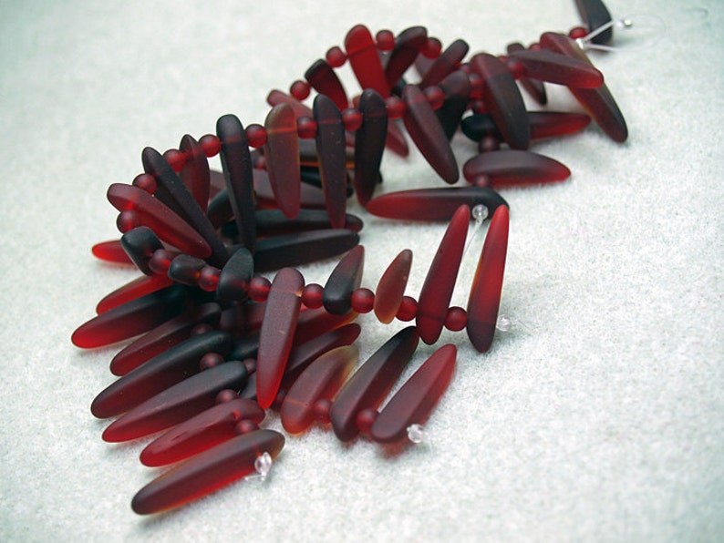 May include: A strand of dark red and amber glass beads in a teardrop shape. The beads are strung on a clear thread and are arranged in a loose, flowing pattern.