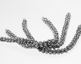 May include: A close-up of a silver beaded chain, with multiple strands of beads twisted together. The beads are small and round, creating a textured look.