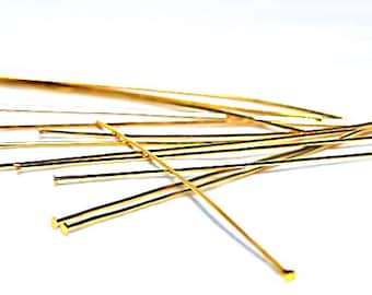 May include: A group of gold-colored head pins, also known as T-pins, are scattered on a white surface. The pins are thin and have a small, round head at one end.