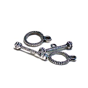 May include: Silver-toned metal clasps with a textured design. The clasps have a round loop and a straight bar with a decorative end.