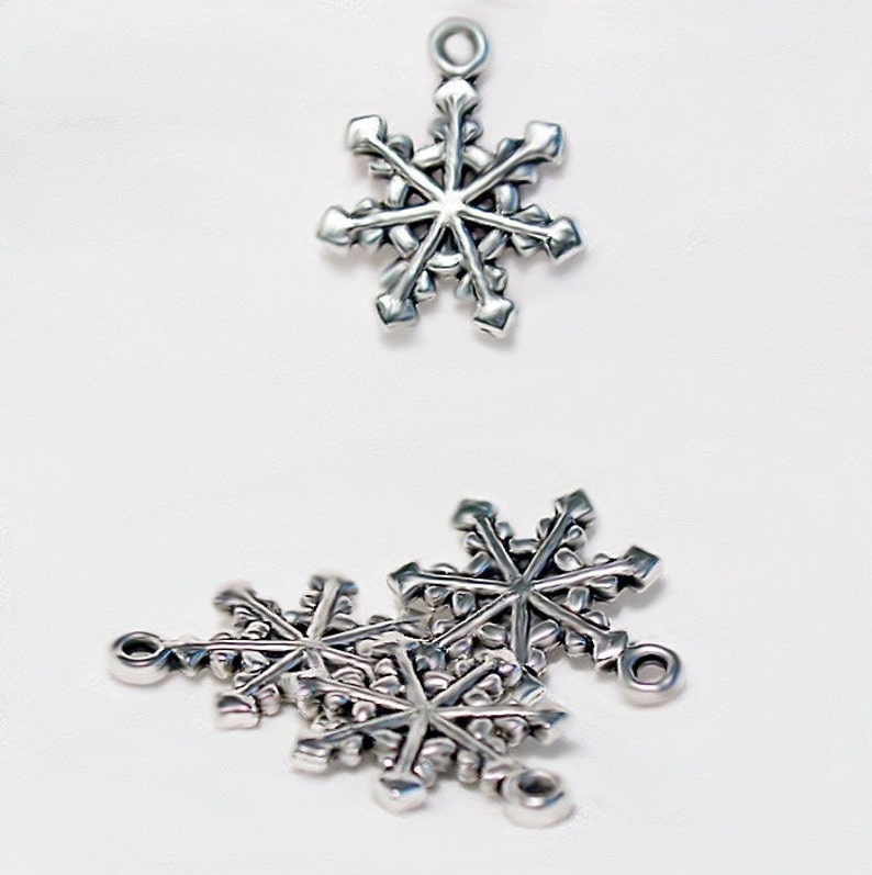Op de afbeelding: Zilveren sneeuwvlokcharms met een lus om op te hangen. De charms liggen in een stapel met &eacute;&eacute;n charm bovenop.