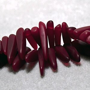 May include: A group of approximately 20 deep red glass beads, each shaped like a long, thin teardrop. The beads are arranged in a loose, overlapping pattern, creating a visually appealing display.