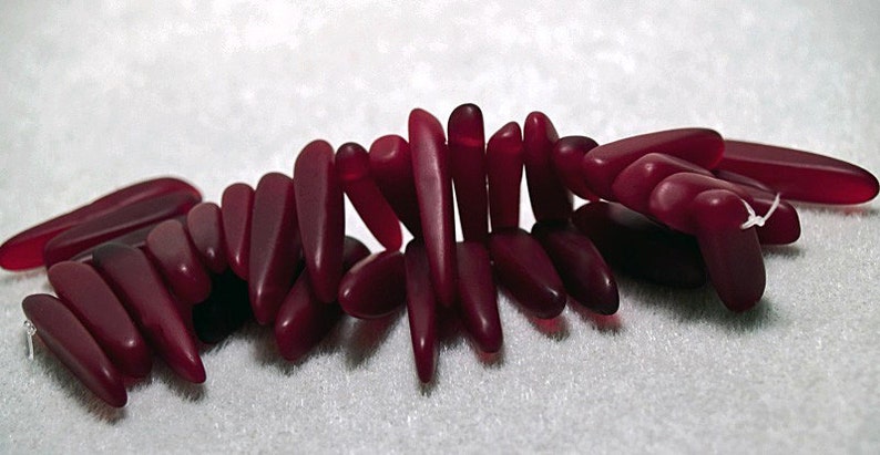 May include: A group of approximately 20 deep red glass beads, each shaped like a long, thin teardrop. The beads are arranged in a loose, overlapping pattern, creating a visually appealing display.