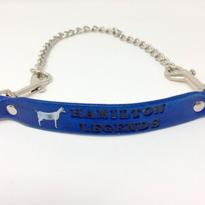 Pu&ograve; includere: Cinturino in pelle blu con una catena argentata e due fibbie. Il cinturino presenta una silhouette di capra bianca e il testo "HAMILTON LEGENDS" in rilievo nero.