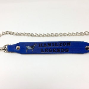 Pu&ograve; includere: Portachiavi in ​​cuoio blu con una catena argentata e un fermaglio argentato. Il cinturino in cuoio presenta una piccola silhouette di capra e il testo "HAMILTON LEGENDS" in rilievo in nero.