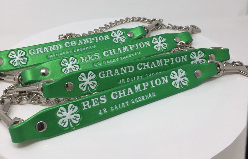 May include: Four green leather straps with silver chains and metal clasps. Each strap has a white four-leaf clover with the letters "H" printed on each leaf. The straps have white text that reads "Grand Champion", "Res Champion", "Grand Champion", and "Res Champion".