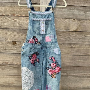 May include: Light blue denim overalls with floral patches and a white lace doily. The overalls feature pink and red rose appliques, and patches of pink, blue, and plaid fabric. The straps are trimmed with white lace.