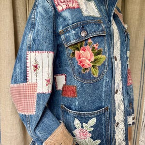 May include: A blue acid-wash denim jacket featuring various fabric patches, including floral and plaid patterns. The jacket has a tan corduroy collar and cuffs, a front button closure, two chest pockets, and white lace trim running vertically along the front.