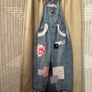 May include: Blue denim overalls featuring a light wash, white paint splatters, and decorative patches. The bib includes white lace trim and three buttons. The pockets have white lace accents, and the legs feature various fabric patches including a pink floral print, striped fabric, and embroidered squares.