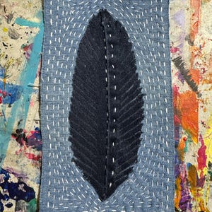 Pode incluir: Uma peça de arte em tecido de ganga com um design de folha azul escuro. A folha é costurada sobre um fundo de ganga azul claro com fio branco. A peça é retangular e tem uma aparência costurada à mão.