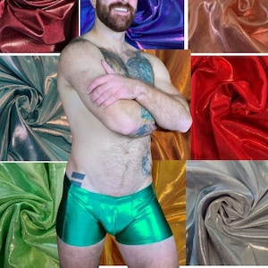 Puede incluir: Un hombre con pantalones cortos metálicos verdes con un acabado brillante. Los pantalones cortos tienen una cintura baja y un ajuste ceñido. El hombre está de pie frente a un fondo de telas de diferentes colores.