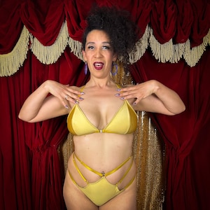 May include: A gold-colored lingerie set featuring a triangle-shaped bra with a center clasp and high-waisted bottoms with multiple strap details. The set is made of a shiny fabric. The background is a red curtain with gold fringe.