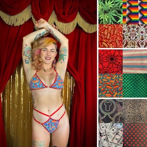May include: A woman wearing a red and blue bikini with a hot dog print. The bikini top is a triangle style and the bottom is a high-waisted style with a criss-cross design. The woman is standing in front of a red velvet curtain with gold fringe.