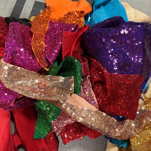 May include: A pile of colorful sequined fabric scraps in various shades of red, pink, purple, green, orange, gold, and blue. The fabric scraps are arranged on a gray surface with a ruler.