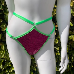 Puede incluir: Una ropa interior de lentejuelas rosa con ribetes y tirantes de color verde neón. La ropa interior tiene forma de diamante con un corte en el centro.
