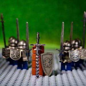 May include: A set of four silver toy knights with swords and shields standing on a white and gray tiled surface. The knights are wearing silver armor and have red and silver helmets. The shields feature a silver lion design. A silver sword with a brown handle is in the center of the image.