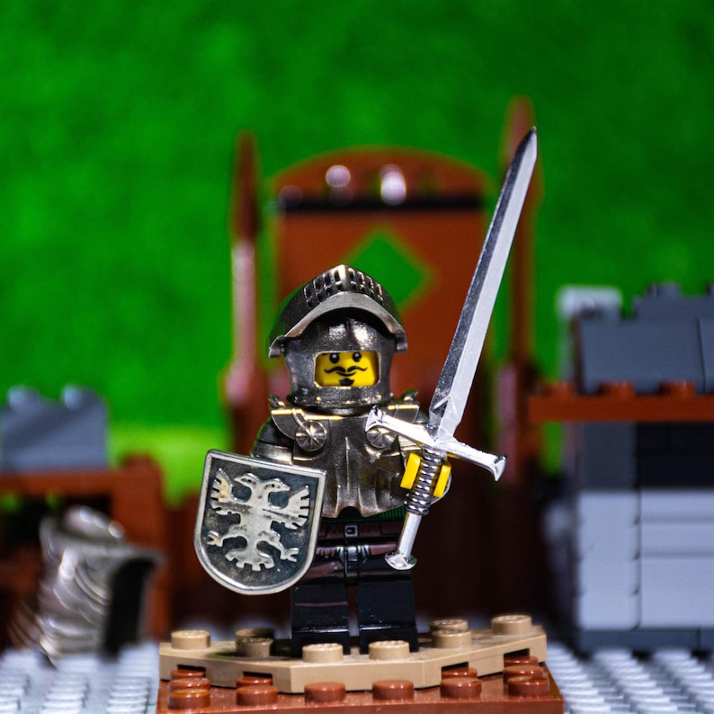 Puede incluir: Una figura de caballero Lego de plata y negro con una cara amarilla, que lleva un casco y una armadura, sosteniendo una espada plateada y un escudo con un dise&ntilde;o de &aacute;guila negro y plateado. El caballero est&aacute; de pie sobre una base marr&oacute;n.