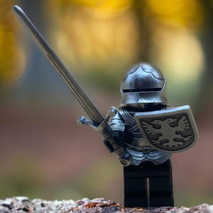 May include: A silver toy knight figure, featuring a helmet, armour, shield, and sword. The shield displays a detailed eagle emblem. The figure is posed as if ready for battle, with the sword held aloft.